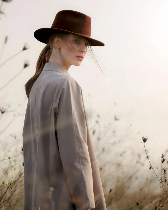 a woman standing in a field wearing a hat, inspired by Henry Bright, wear's beige shirt, back - lit, wearing hunter coat, promo image