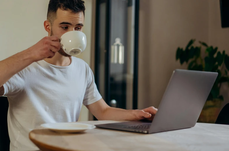 a man sitting at a table with a laptop and a cup of coffee, a screenshot, pexels contest winner, happening, lachlan bailey, young male, is ((drinking a cup of tea)), maintenance