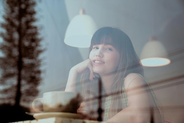 a woman sitting at a table with a cup of coffee, profile image, hazy, looking through a window frame, smiling slightly