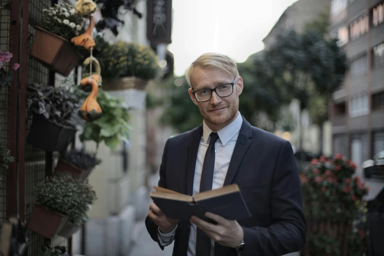 a man in a suit and tie reading a book, a portrait, by Emma Andijewska, pexels contest winner, standing in a city street, one man is blond, nerdy appearance, avatar image