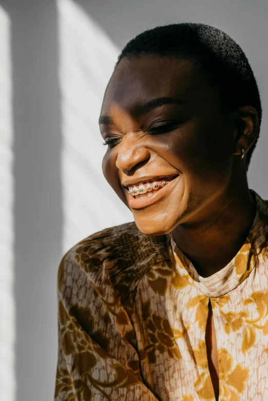 a smiling woman sitting in front of a window, trending on unsplash, brown skin. light makeup, gold dappled light, head bent back in laughter, patterned clothing