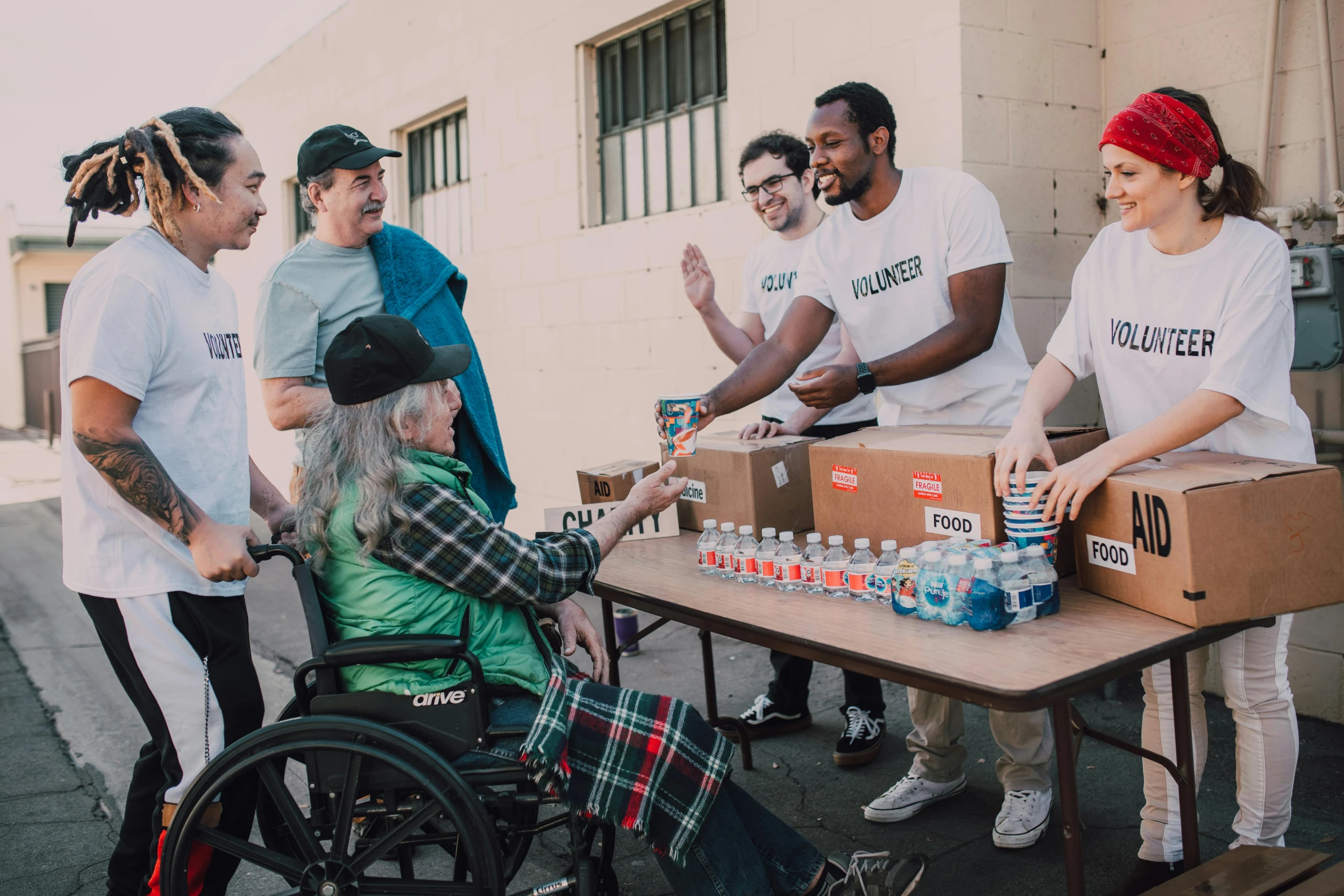 a group of people standing around a table, by Dan Frazier, pexels contest winner, process art, sitting in a wheelchair, bay area, giving gifts to people, hunger