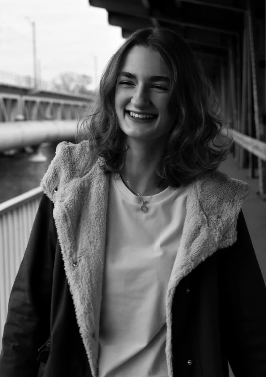 a black and white photo of a woman smiling, a black and white photo, by Felix-Kelly, girl wearing hoodie, on a bridge, wavy shoulder-length hair, medium format. soft light