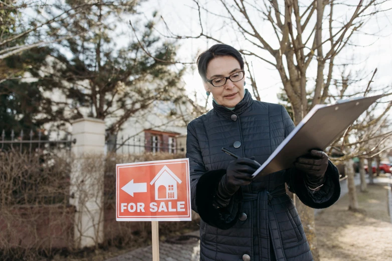a woman standing in front of a for sale sign, by Emma Andijewska, shutterstock, writing on a clipboard, standing outside a house, sangsoo jeong, he is holding a large book