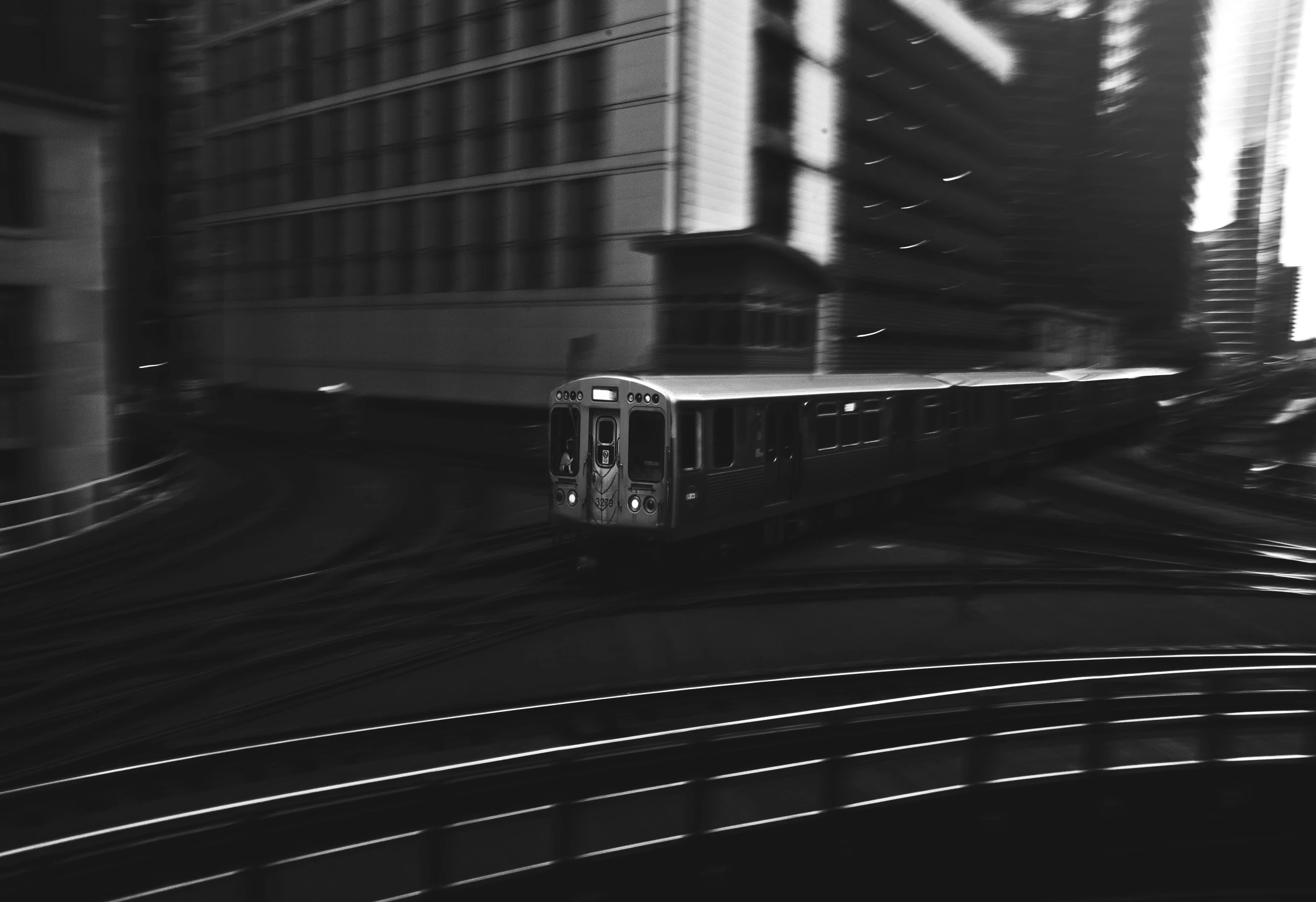 a train traveling through a city next to tall buildings, a black and white photo, unsplash, low quality footage, nanae kawahara, motion photo, consist of shadow