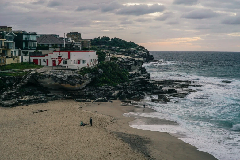 a couple of people standing on top of a sandy beach, a matte painting, pexels contest winner, bondi beach in the background, the village on the cliff, overcast dawn, image from afar