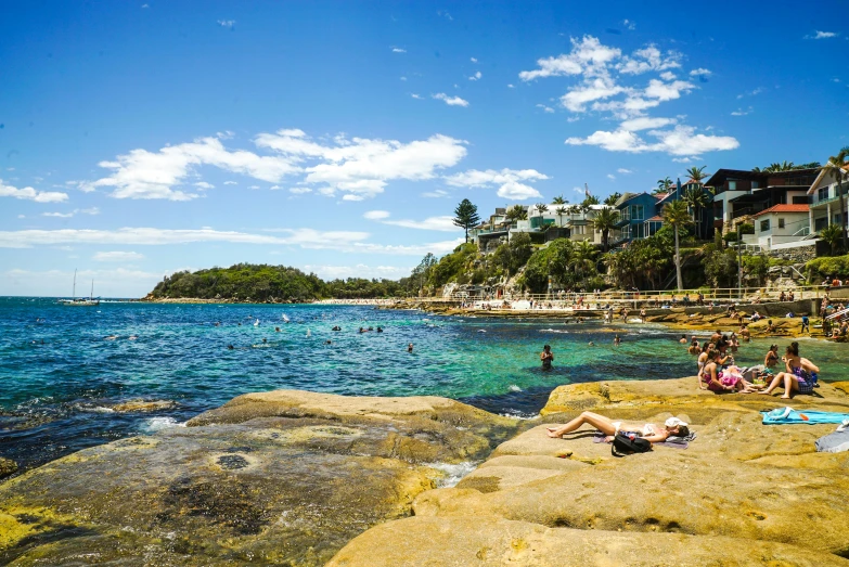 a group of people sitting on top of a rock next to the ocean, manly, crystal clear blue water, people swimming, a quaint