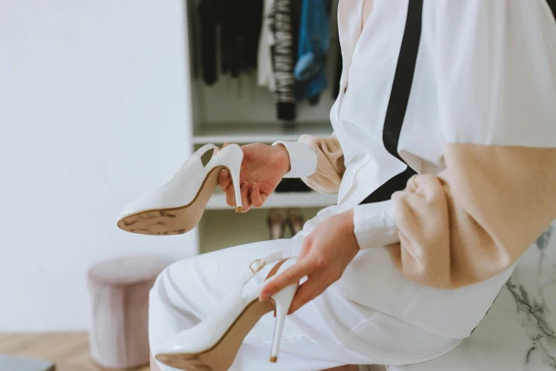 a woman sitting on top of a bed holding a pair of shoes, trending on pexels, white suit and black tie, sitting on a mocha-colored table, talons, white coat