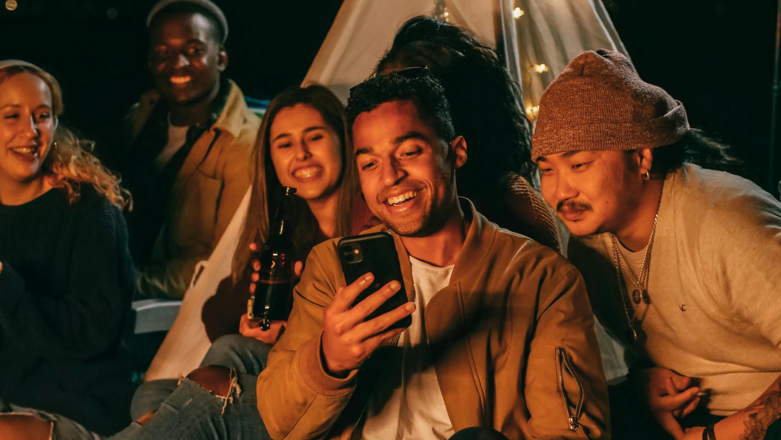 a group of people sitting around a campfire looking at a cell phone, a portrait, trending on pexels, realism, brown skin man with a giant grin, nvidia promotional image, rooftop party, holding a very advance phone