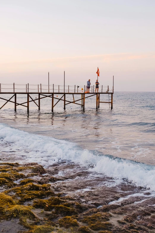 a man standing on top of a pier next to the ocean, agrigento, people angling at the edge, serene beach setting, dusk setting