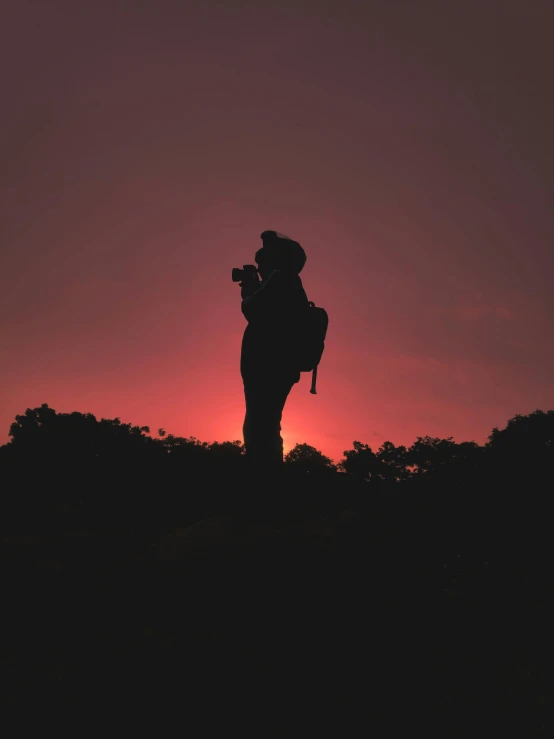 a person standing on top of a hill at sunset, holding a camera, profile picture, the sky is a faint misty red hue, photograph captured in a forest