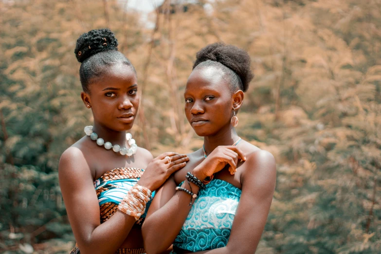 a couple of women standing next to each other, by Chinwe Chukwuogo-Roy, trending on pexels, afrofuturism, tribal jewelry, black teenage girl, bare shoulders, brown and cyan color scheme