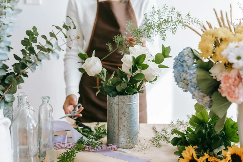 a woman standing in front of a table filled with vases of flowers, trending on pexels, arts and crafts movement, grey and blue theme, insisted on cutting in line, ingredients on the table, bouquet