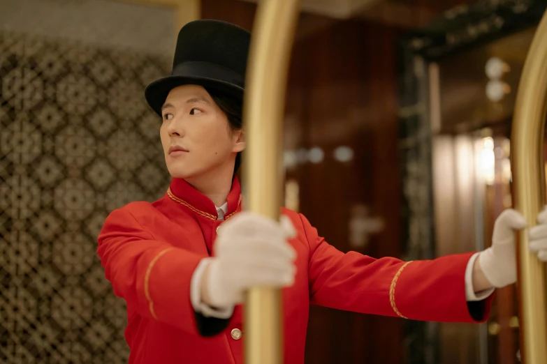 a man in a red jacket and white gloves, pexels contest winner, she is holding a long staff, hotel room, wearing cylinder hat, maggie cheung