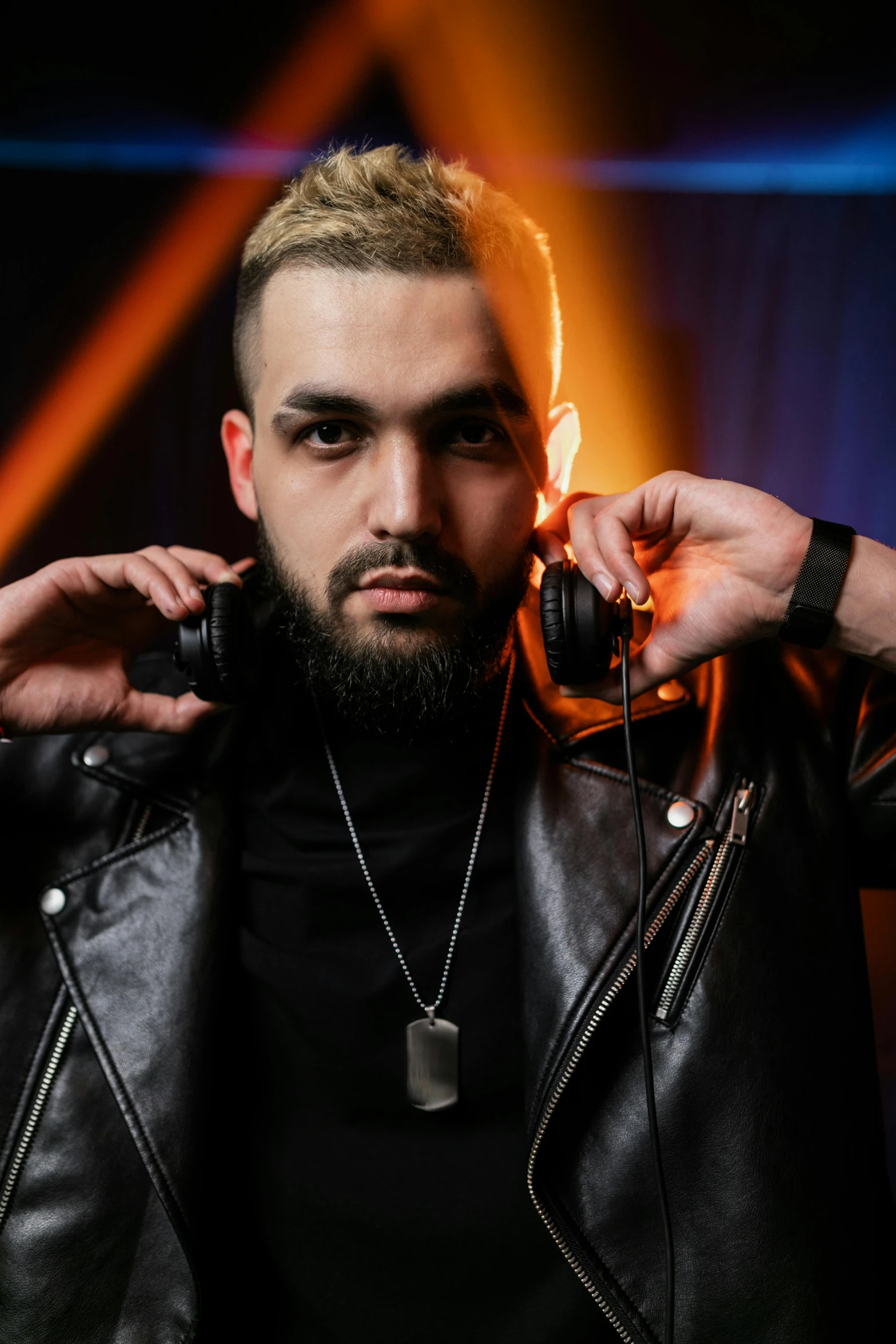 a man in a leather jacket holding a pair of headphones, inspired by Volkan Baga, dark lighting, light beard, riyahd cassiem, dj at a party