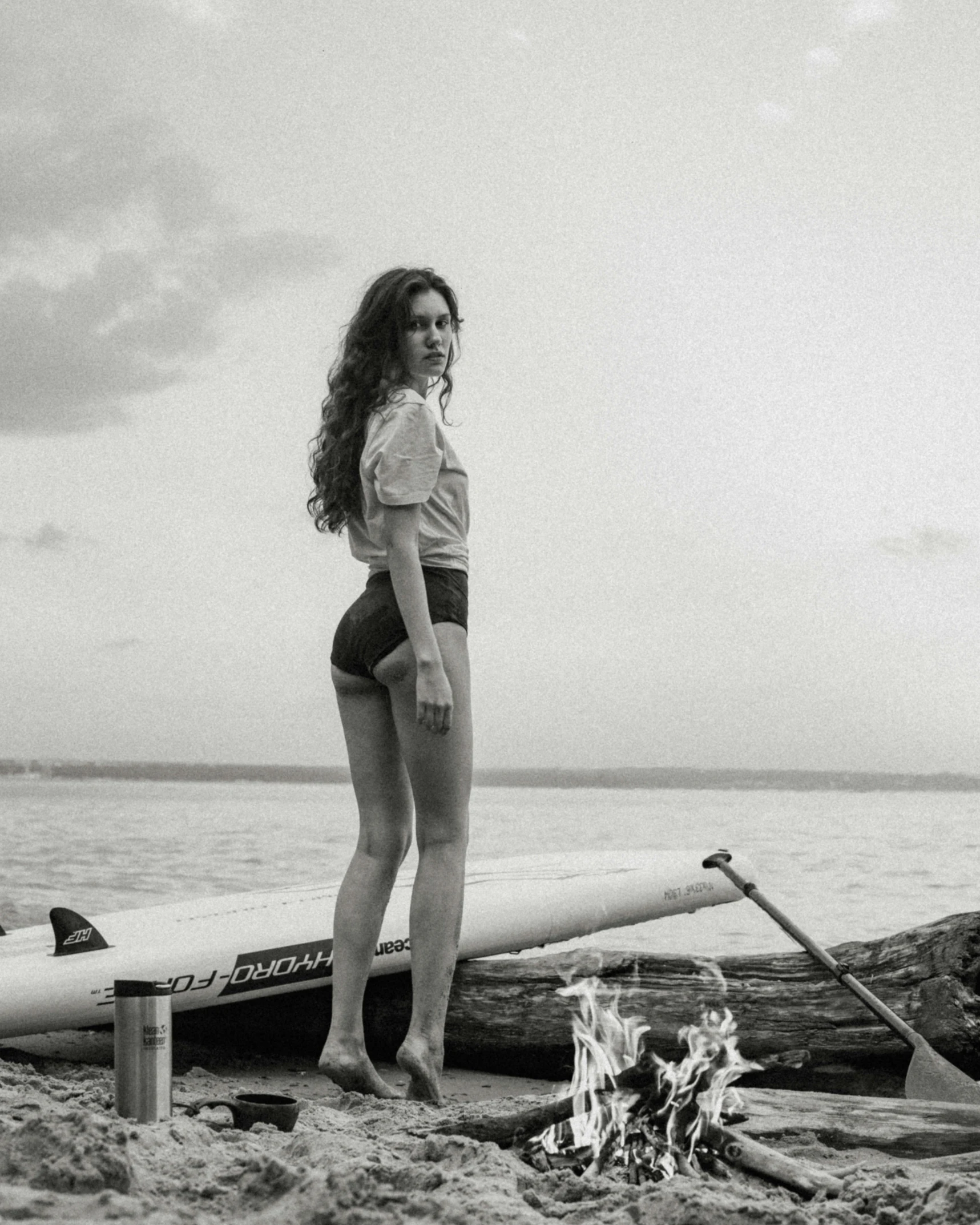 a woman standing on a beach next to a surfboard, a black and white photo, renaissance, on a boat, gal gadot, non binary model, alternate album cover