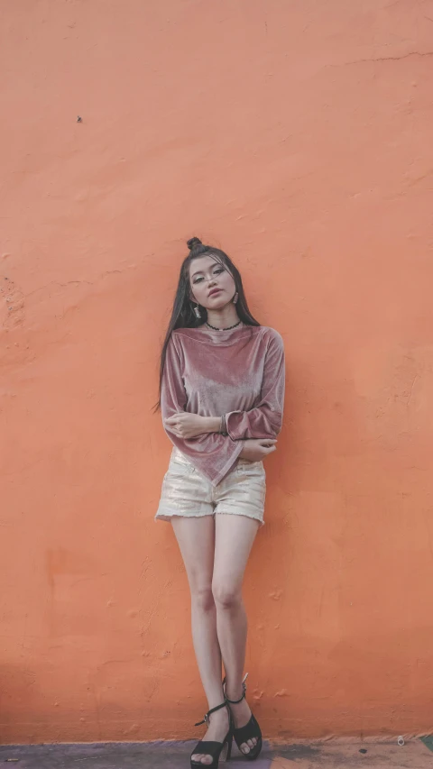 a woman standing in front of an orange wall, inspired by Elsa Bleda, pexels contest winner, aestheticism, croptop and shorts, long sleeves, portrait of modern darna, faded pink