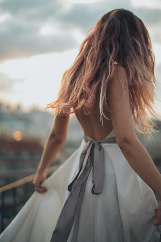 a woman in a white dress walking across a bridge, trending on unsplash, long flowing pink hair, hands behind back, standing on a rooftop, ombre velvet gown