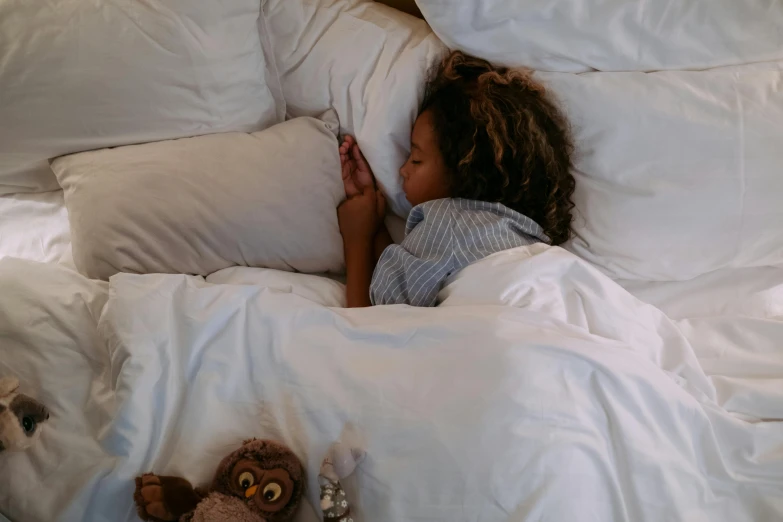 a little girl laying in bed with a stuffed animal, by Elsa Bleda, pexels contest winner, happening, with brown skin, with an owl on her shoulder, white pillows, high angle