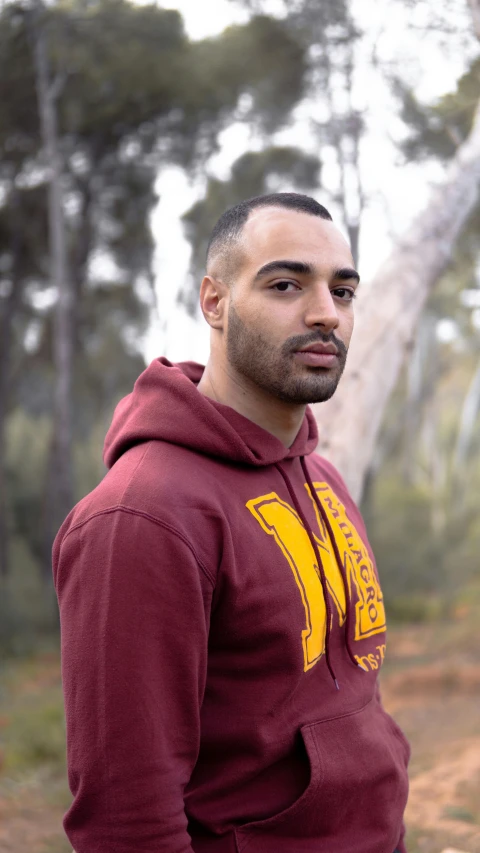 a man in a maroon hoodie standing in a forest, aboriginal australian hipster, joe taslim, lgbtq, official photo