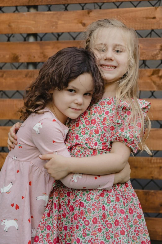 two little girls hugging each other in front of a fence, dressed in a flower dress, full product shot, modeled, f / 2 0