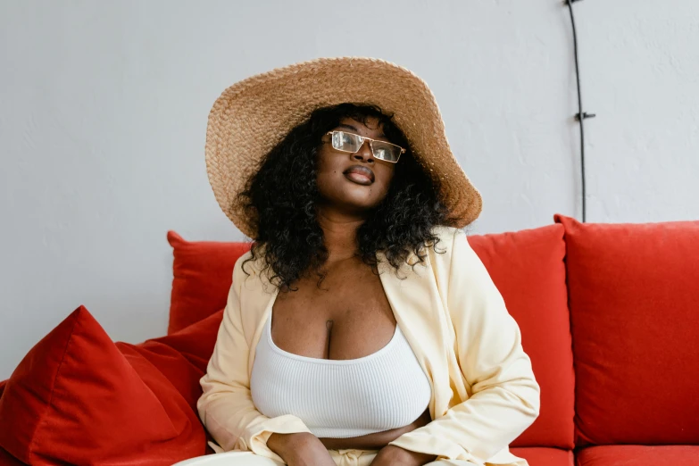 a woman sitting on a red couch wearing a hat, pexels contest winner, brown skin, plus-sized, revealing outfit, square rimmed glasses