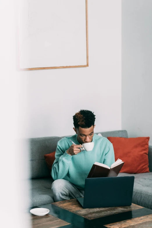 a man sitting on a couch using a laptop, trending on pexels, happening, with teal clothes, reading a book, next to a cup, ashteroth