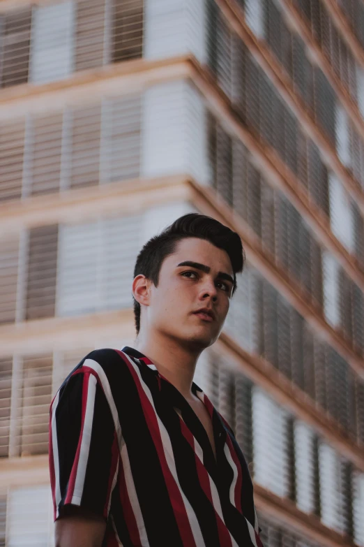 a man standing in front of a tall building, an album cover, by Alejandro Obregón, pexels contest winner, wearing stripe shirt, thick jawline, male teenager, headshot profile picture