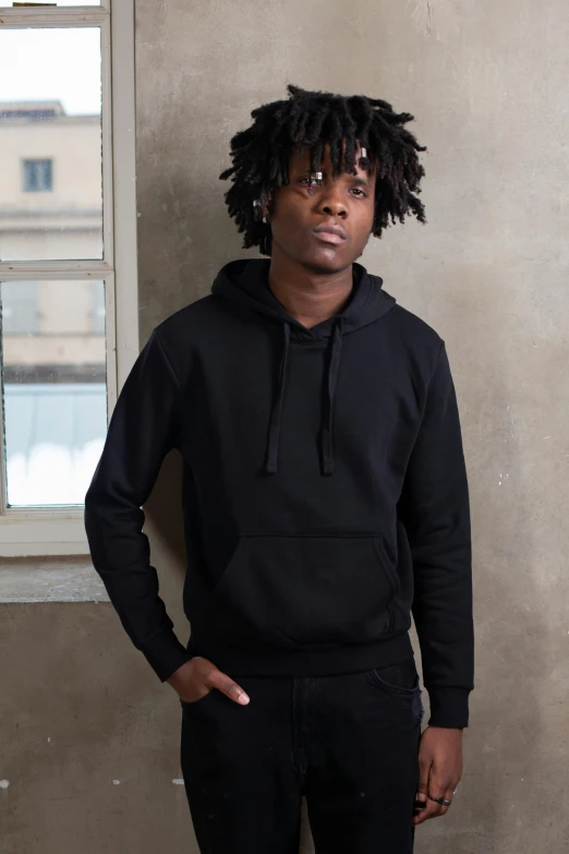 a man with dreadlocks standing in front of a window, wearing a black hoodie, main colour - black, hero shot, tailored clothing