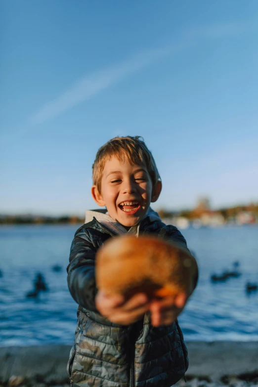 a young boy holding a donut in front of a body of water, laughing, unsplash photo contest winner, subject= duck, holding a skull