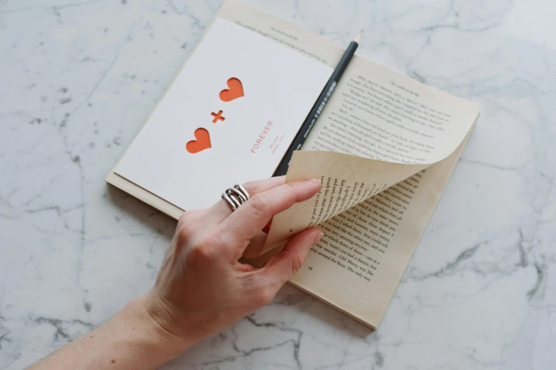 a person holding a pen over an open book, an album cover, by Emma Andijewska, pexels contest winner, private press, reading the book about love, pencil marks, storybook design, on a canva