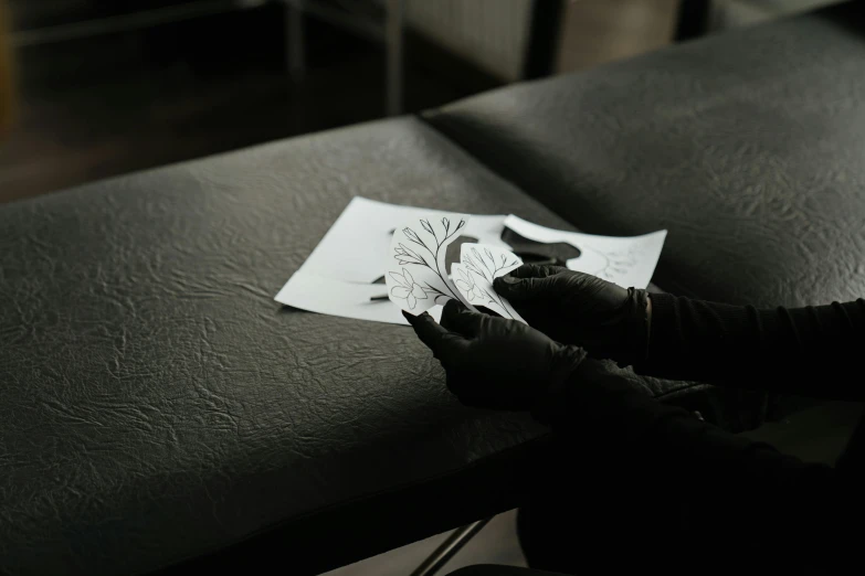 a person is drawing on a piece of paper, an ink drawing, by Adam Marczyński, pexels contest winner, black bandage on arms, sitting in a lounge, paper cut art, tattoo