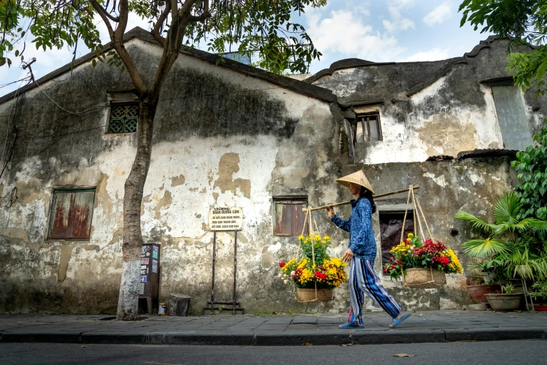 a woman walking down a street past a building, inspired by Cui Bai, pexels contest winner, flowers around, thatched roofs, style steve mccurry, on center