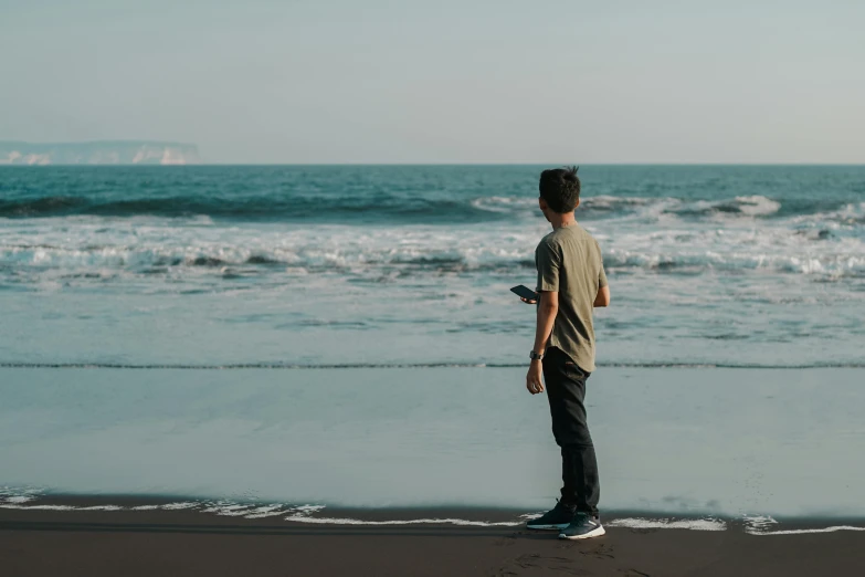 a man standing on top of a beach next to the ocean, pexels contest winner, happening, looking at his phone, slightly turned to the right, simple stylized, thin young male
