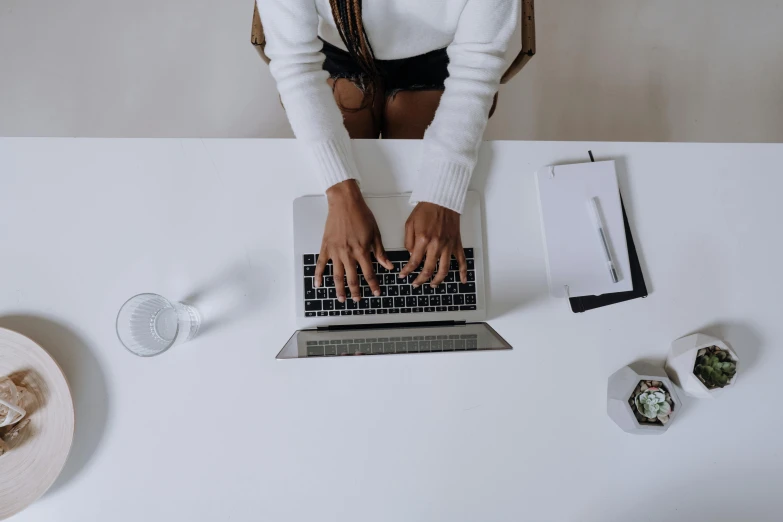 a woman sitting at a table typing on a laptop, by Carey Morris, trending on unsplash, wearing a white sweater, shot from above, standing on a desk, intricate writing