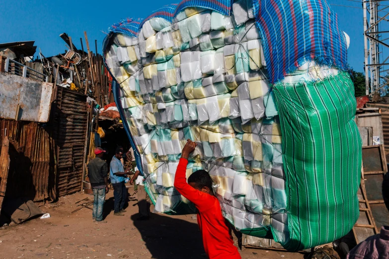 a man standing next to a pile of plastic bottles, an album cover, by Ingrida Kadaka, pexels contest winner, arte povera, weaving, low - angle shot from behind, fabrics, madagascar