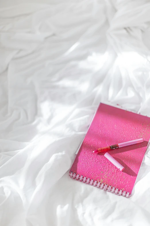 a pink notebook sitting on top of a white bed, lipstick, ((pink))