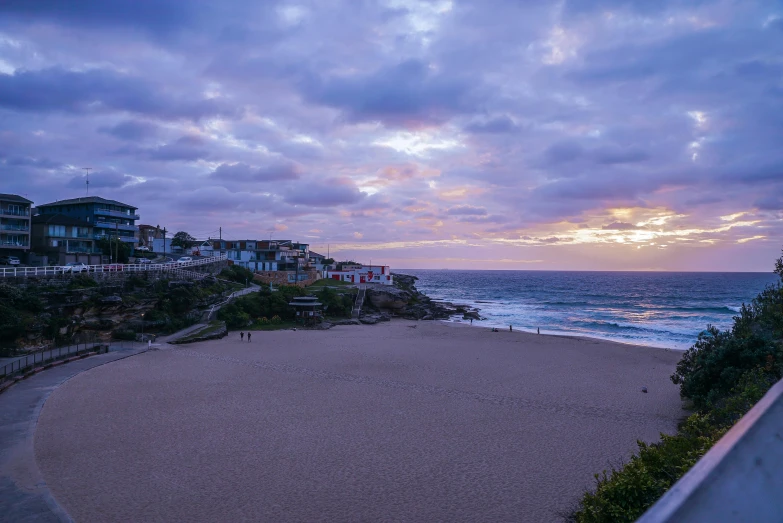 a sandy beach next to the ocean under a cloudy sky, by Elsa Bleda, pexels contest winner, vista of a city at sunset, cliffside town, manly, violet and yellow sunset