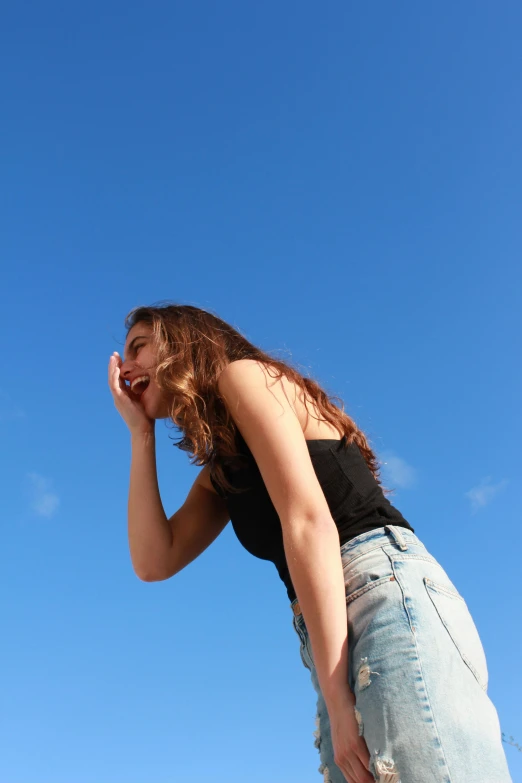 a woman standing on a beach talking on a cell phone, trending on pexels, happening, blue sky above, wearing a black cropped tank top, giggling, seen from below