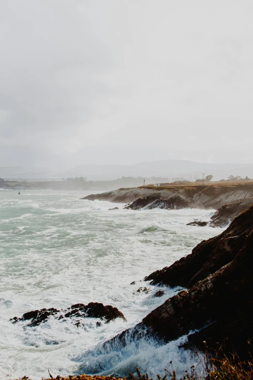 a man standing on top of a cliff next to the ocean, by Robbie Trevino, unsplash, stormy overcast, bay area, today\'s featured photograph 4k, turbulent waves