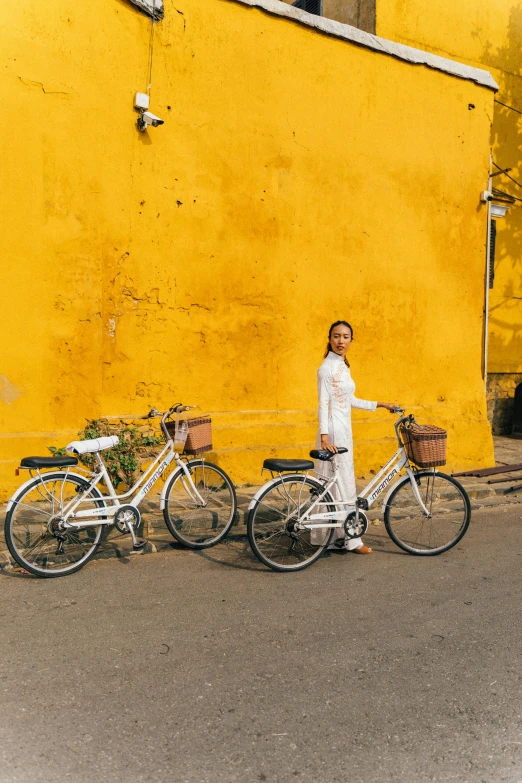 a woman standing next to two bikes in front of a yellow building, pexels contest winner, white and gold color scheme, vietnam, linen, promotional image