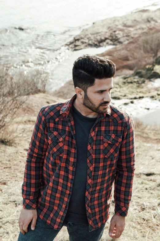 a man standing in a field next to a body of water, an album cover, inspired by Nadim Karam, pexels contest winner, wearing plaid shirt, light stubble with red shirt, caramel. rugged, john redcorn