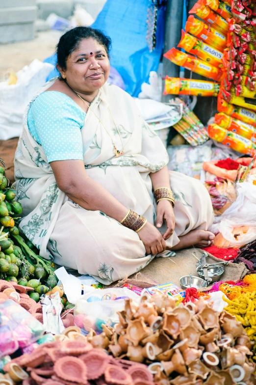 a woman sitting in front of a pile of food, by Ingrida Kadaka, trending on unsplash, hindu ornaments, old lady cyborg merchant, square, market setting
