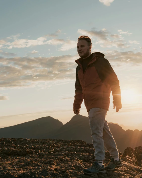 a man standing on top of a mountain at sunset, an album cover, inspired by Hallsteinn Sigurðsson, trending on unsplash, happening, model wears a puffer jacket, walking confidently, lgbtq, lachlan bailey