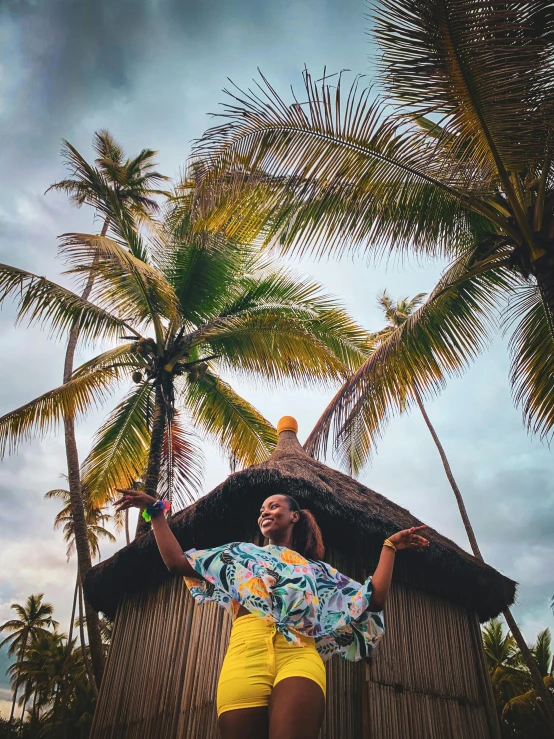 a woman standing in front of a hut with palm trees, by Olivia Peguero, pexels contest winner, pose(arms up + happy), african queen, colorful clothing, profile image