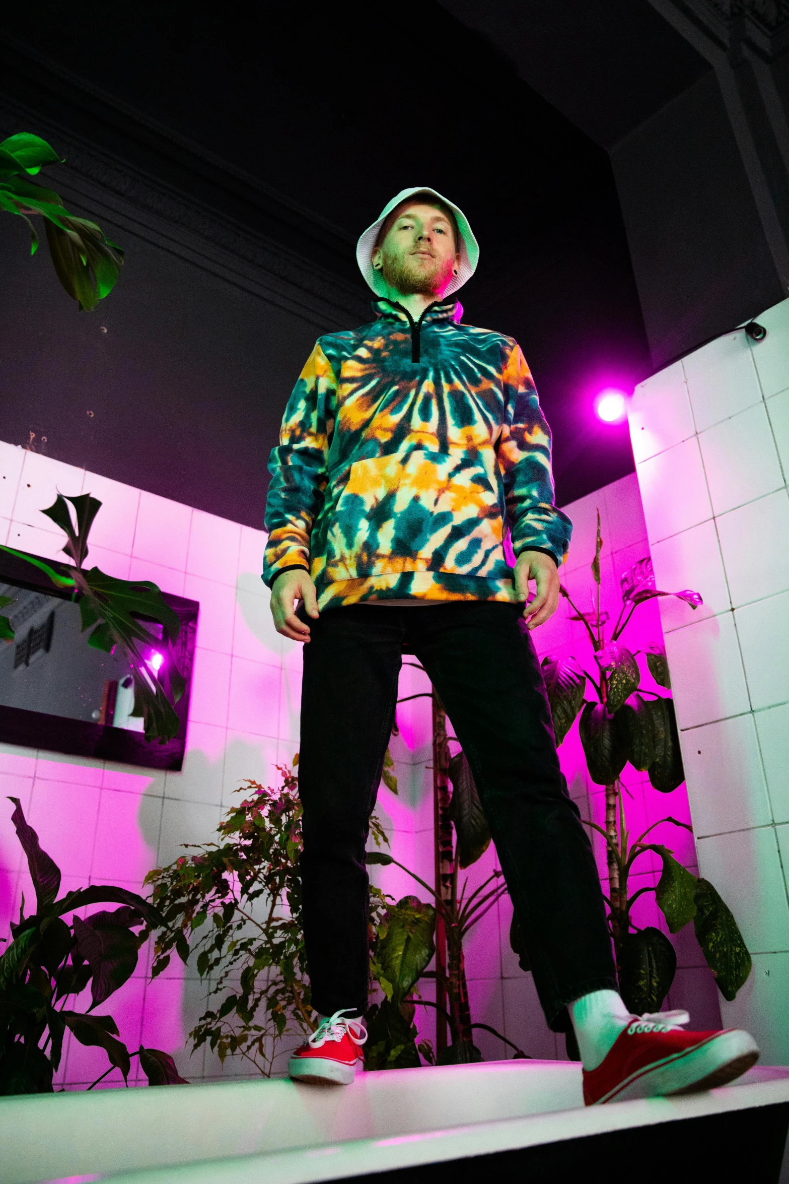 a man standing on top of a white platform, an album cover, unsplash, maximalism, wearing a hoodie, wearing a tie-dye shirt, standing in a dimly lit room, psychedelic vegetation