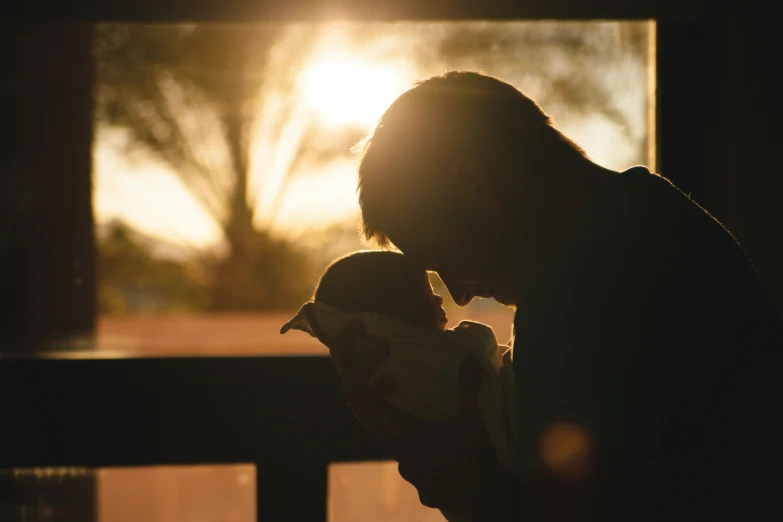 a woman holding a baby in front of a window, pexels contest winner, symbolism, sun behind him, daddy/fatherly, paul barson, silhouette of a man