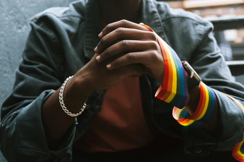 a close up of a person holding a rainbow bracelet, a photo, by Nina Hamnett, trending on pexels, renaissance, androgynous male, dark-skinned, white ribbon, where a large