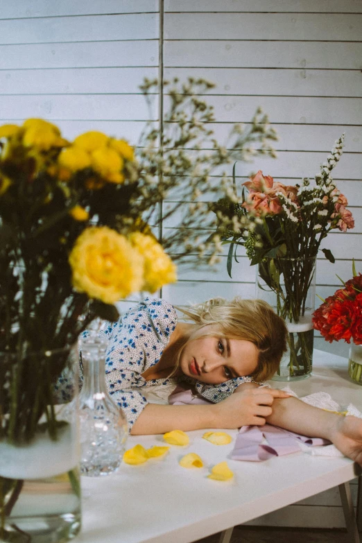 a woman laying on a table next to a vase of flowers, trending on pexels, portrait of kim petras, feels good man, britt marling style, wearing yellow floral blouse