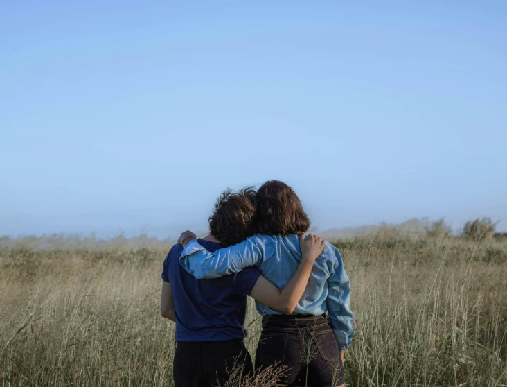 a couple of people that are standing in the grass, by Jessie Algie, unsplash, happening, lesbian embrace, looking onto the horizon, hammershøi, hug